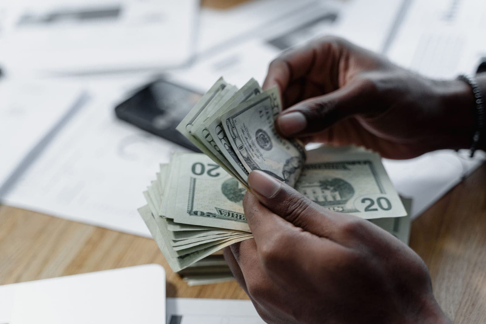 Hands counting US dollar bills on a wooden table beside documents and a phone